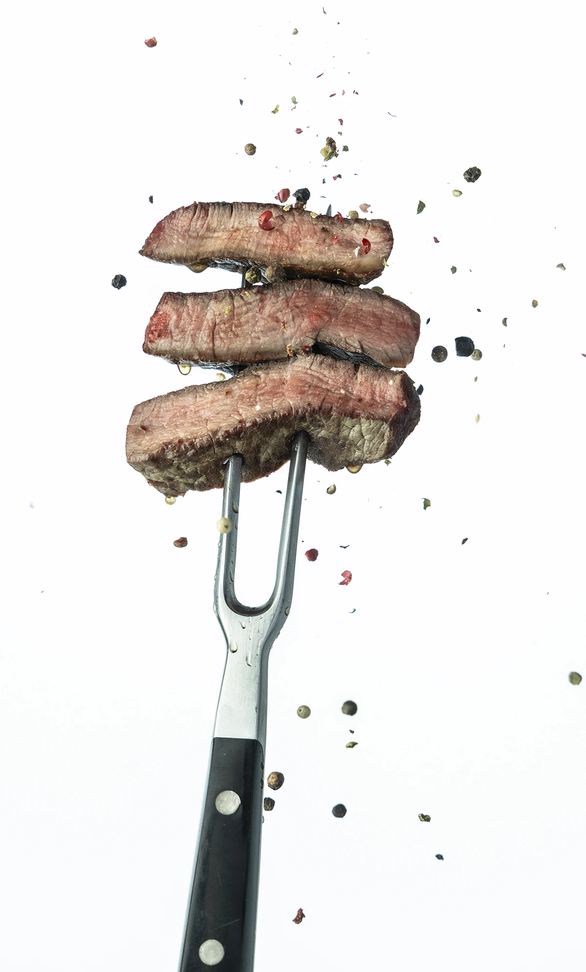 Drei Stücke saftiges Steakstreifen auf einer Gabel mit Pfefferkörnern im Hintergrund.